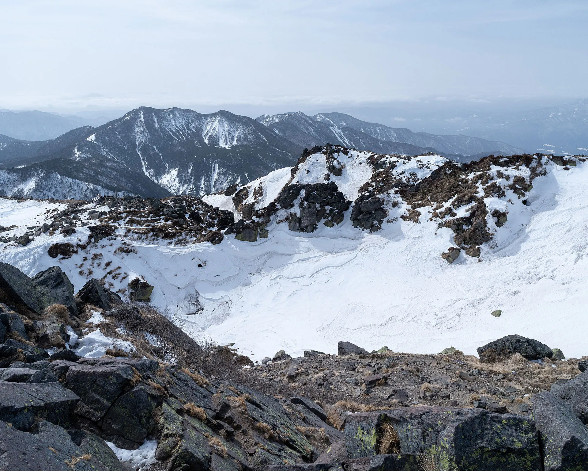 日光白根山の山頂付近から見下ろす巨大な火口跡（すり鉢状の地形）と残雪。岩場と雪のコントラスト。