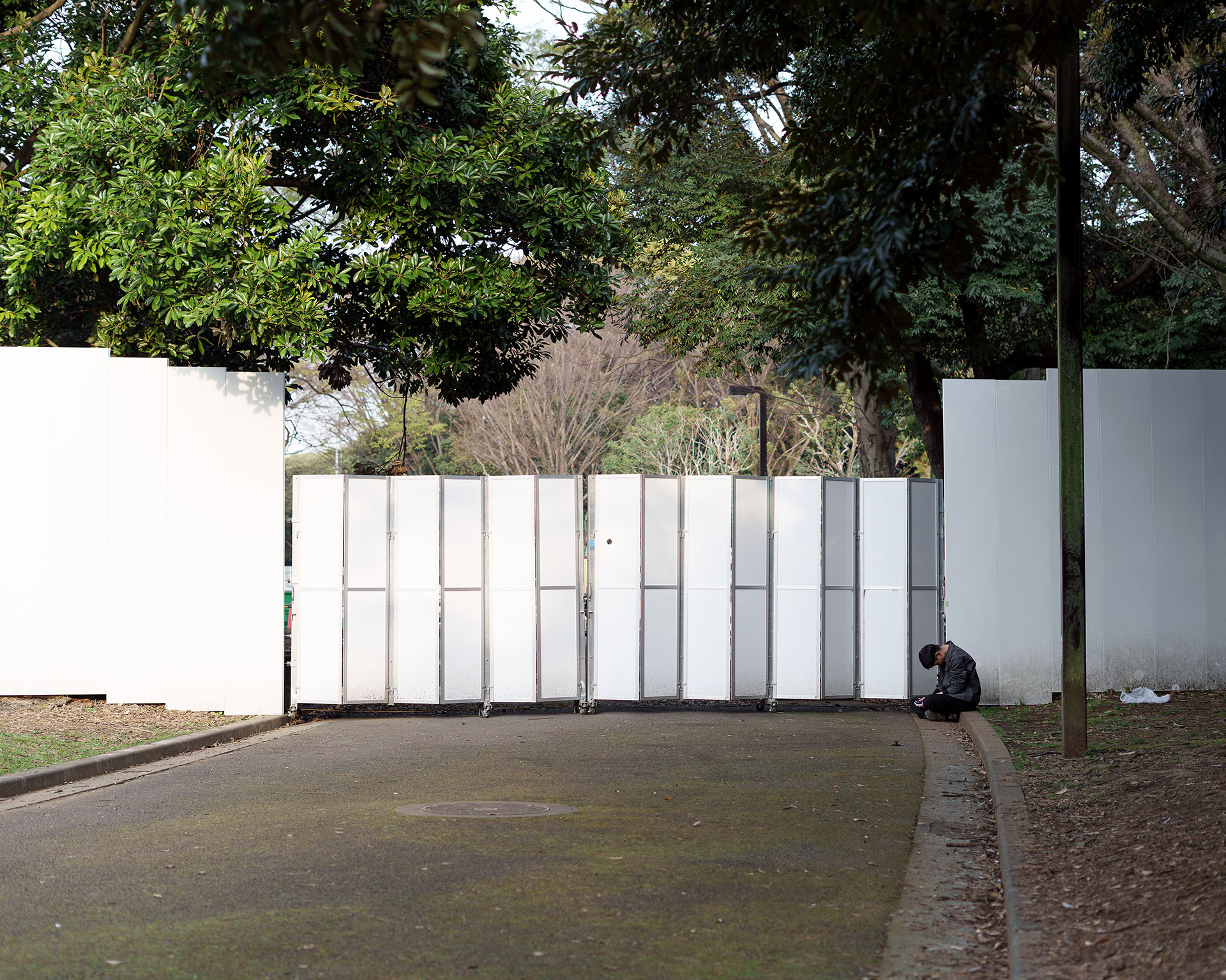 代々木公園内に設置された白い仮囲いゲートの傍らで、静かに座って休憩する一人の男性。