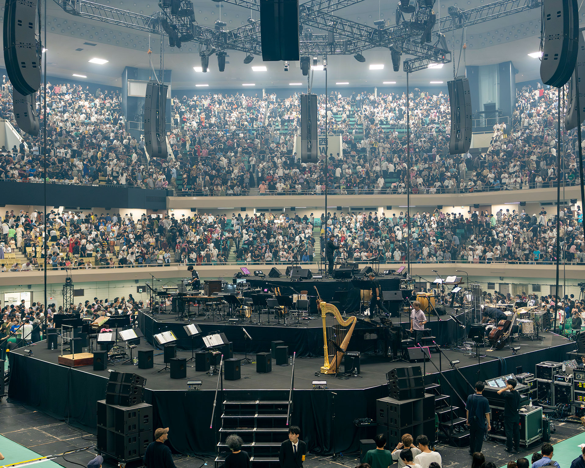 満員の小沢健二「LIFE再現ライブ」日本武道館のコンサート会場内部の様子。大きなステージ上にはハープやその他の楽器、音響機器が設置され、オーケストラのセッティングが整っている。二層に分かれた観客席は多くの観客で埋め尽くされており、天井にはライトやスピーカーなどの機材が設置されている。