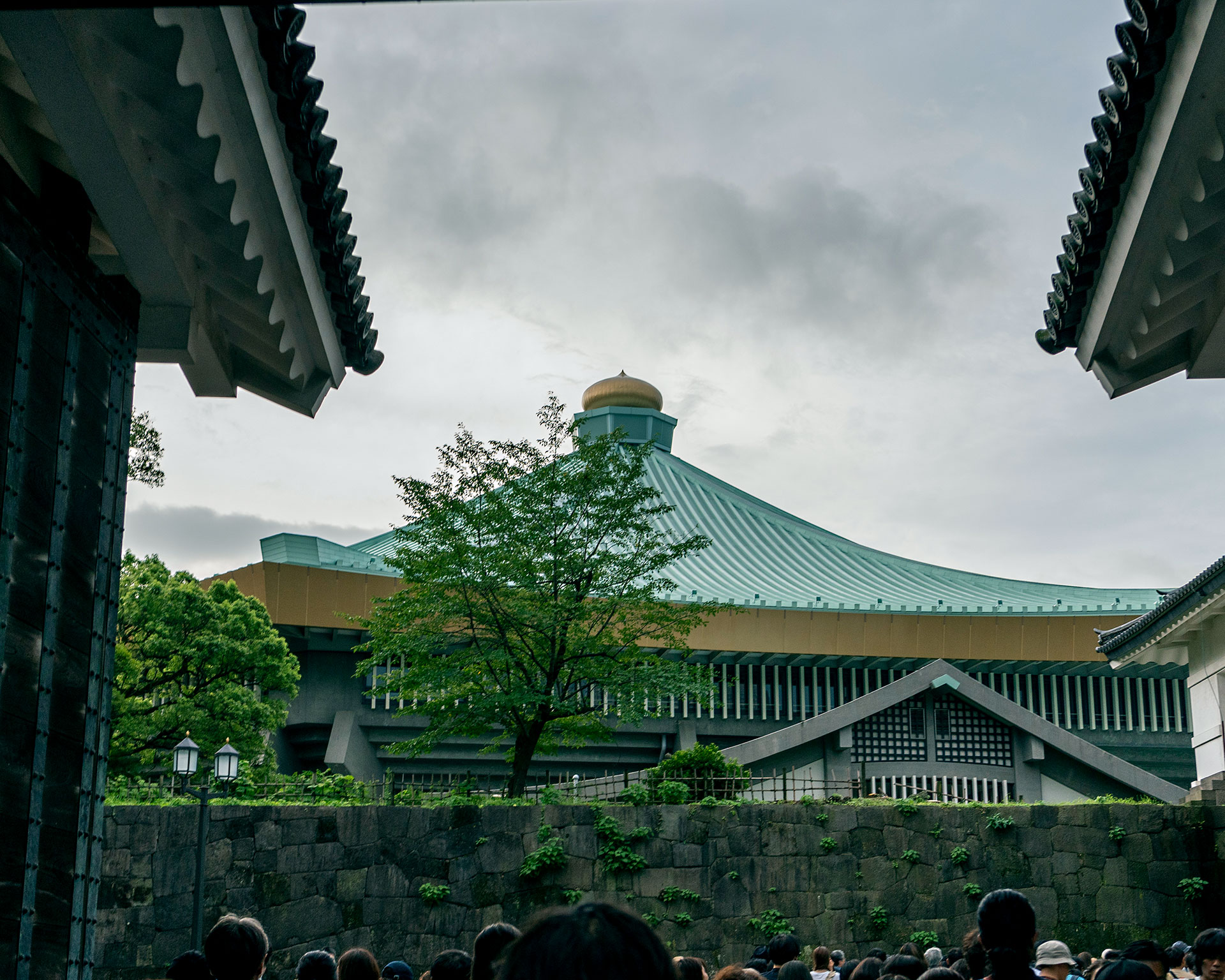 日本武道館の外観を石垣と瓦屋根の間から見上げた構図。特徴的な八角形の建物は緑青色の屋根と金色のドームを持ち、周囲には緑豊かな木々が植えられている。手前には観客の列が見える。伝統的な和風建築の要素とモダンな建築様式が融合した武道館の独特な建築美が捉えられている。