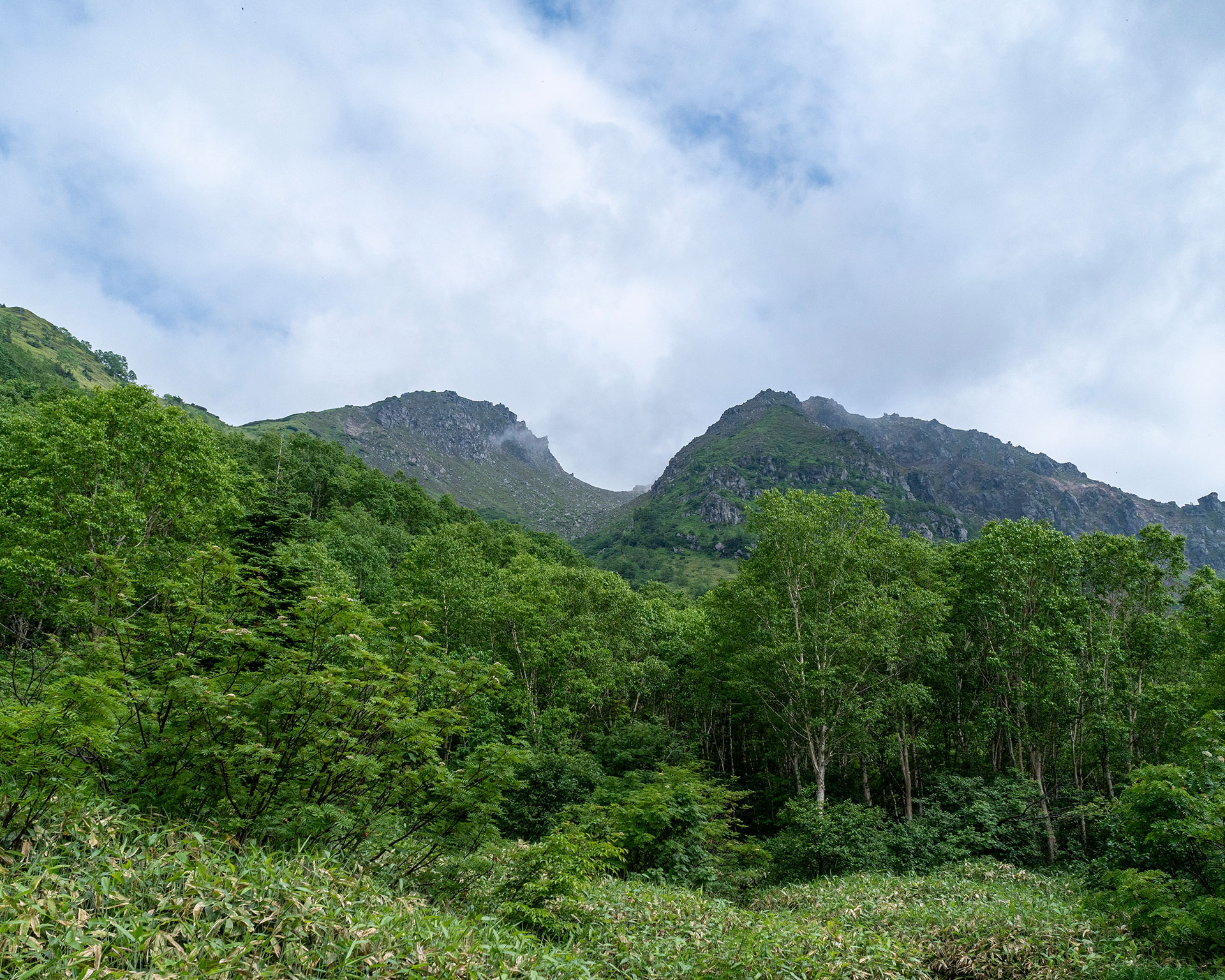 広場から見る緑豊かな山岳風景と雲に覆われた山頂への登山道