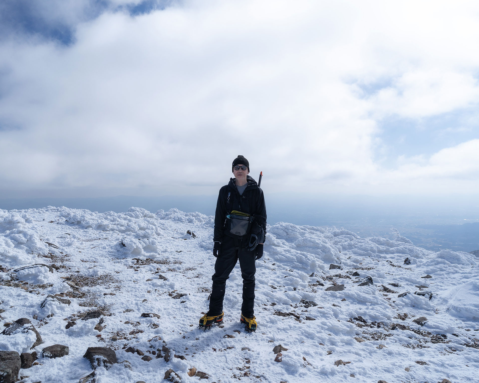 黒いウェアの登山者が雪の山頂に立つ、背景に雲と平野
