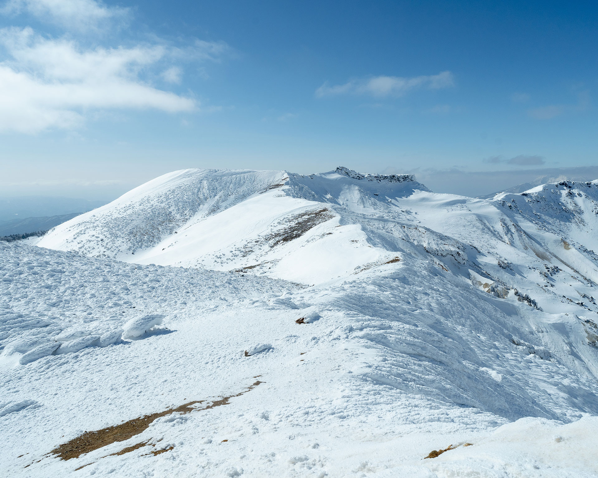 なだらかな雪の稜線が続く山頂部、遠方に山並み