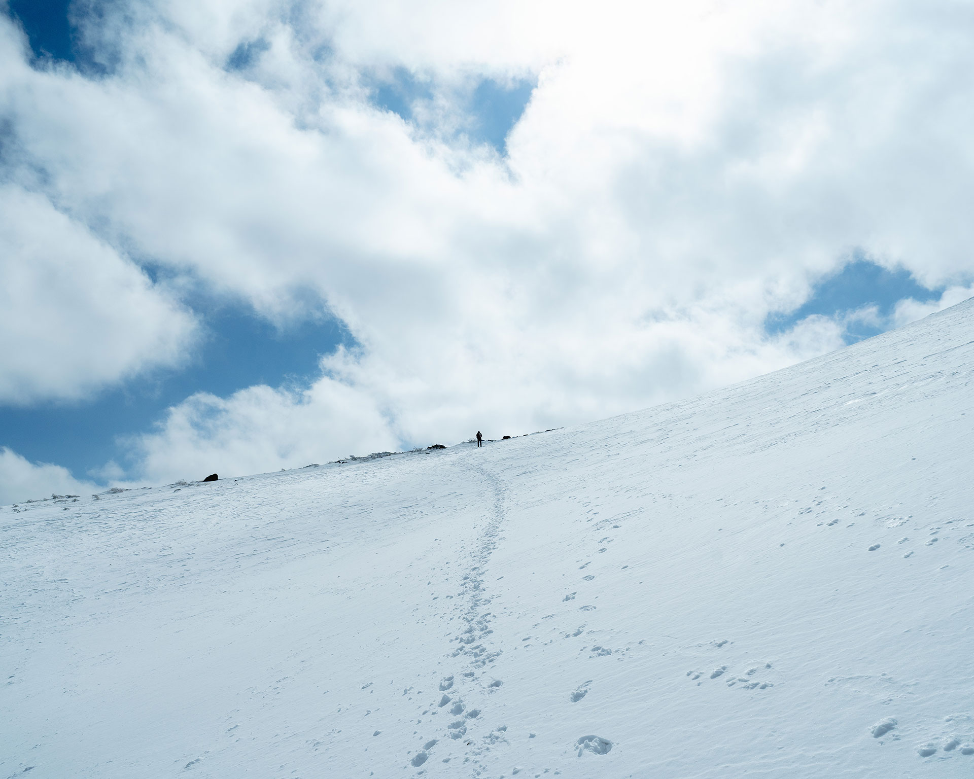 足跡が続く雪の斜面を登る登山者、雲が流れる空