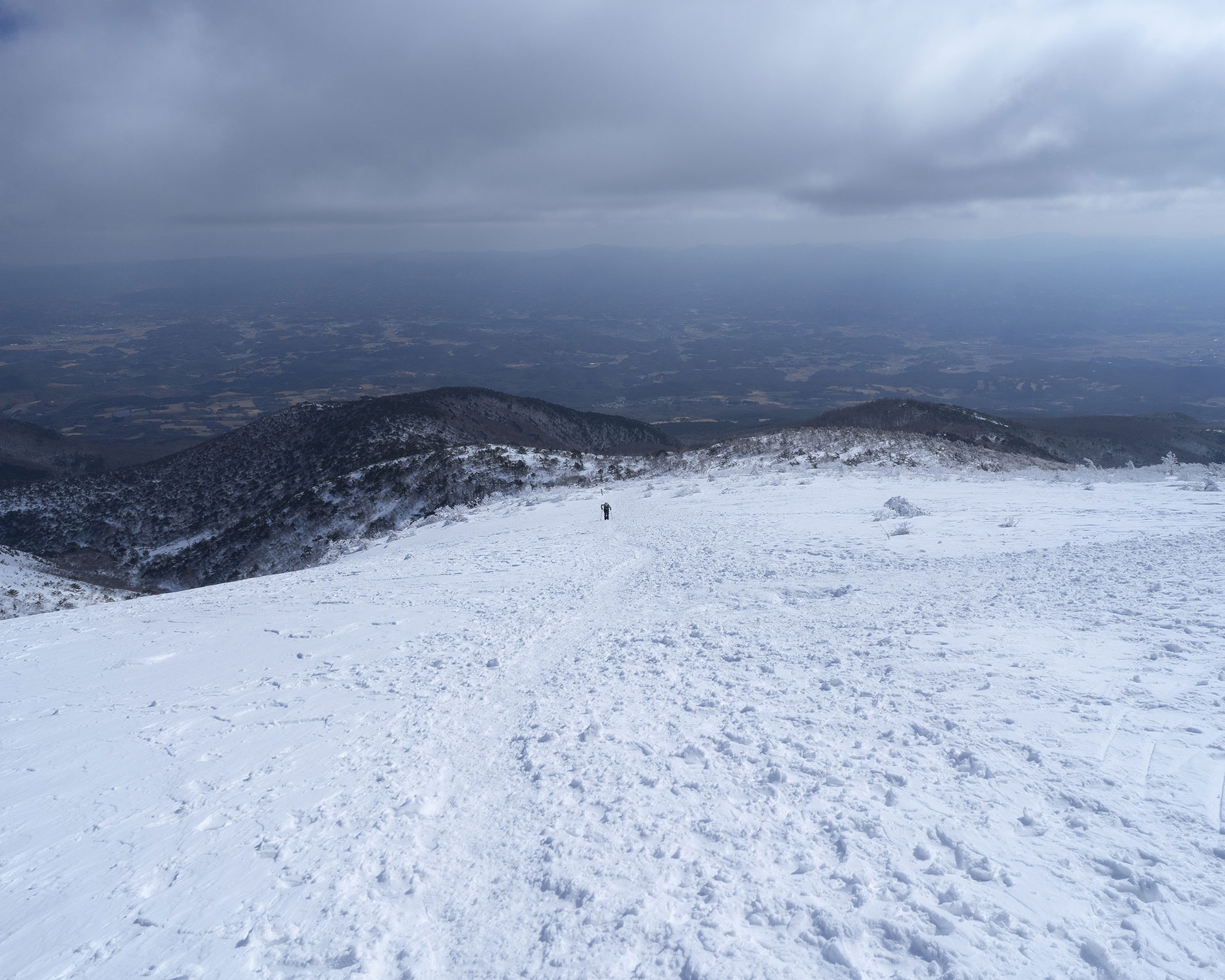 曇り空の下、雪の斜面を登る1人の登山者、遠景に平野部