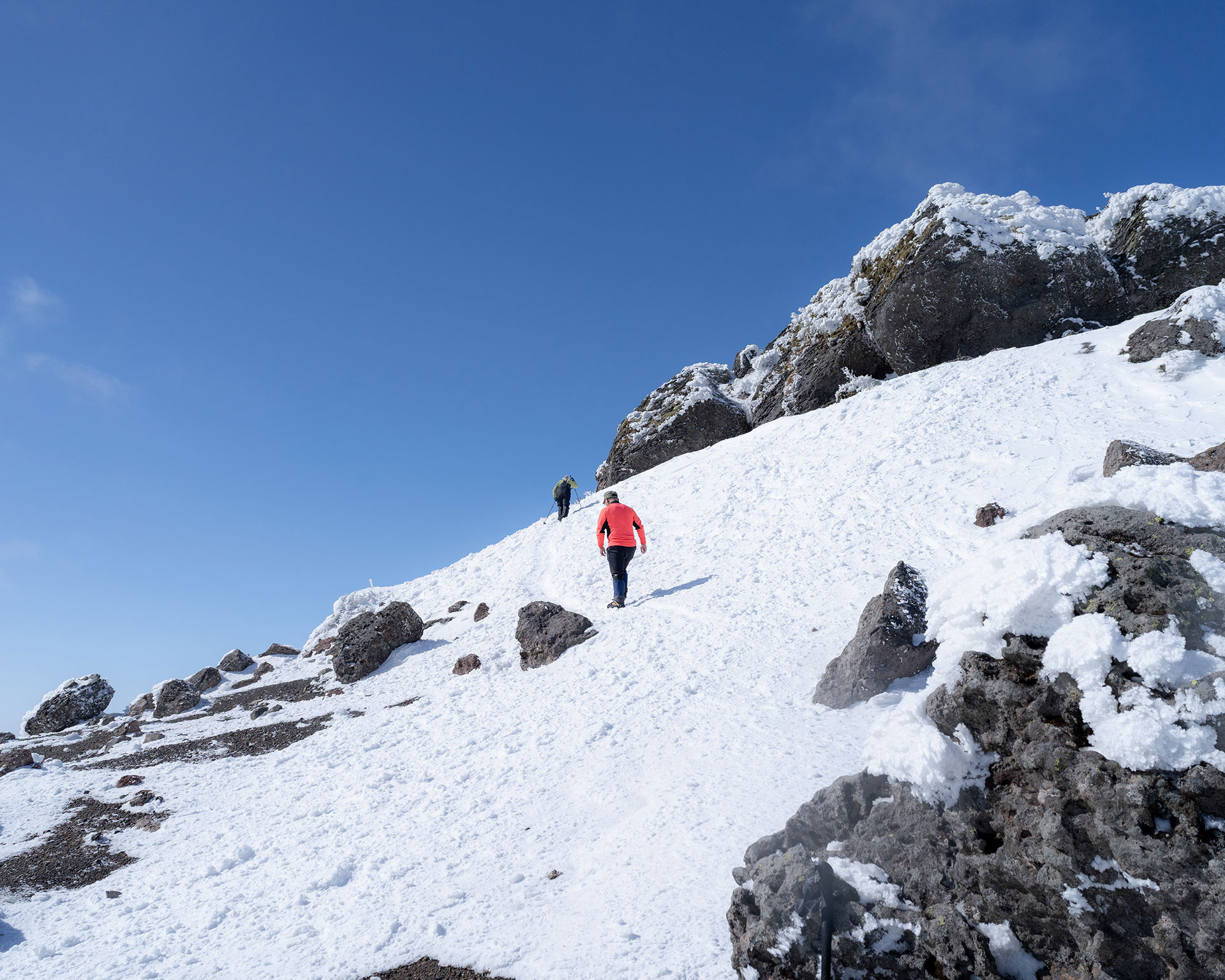 安達太良山の山頂へと岩場の急斜面を登る赤いジャケットの登山者、雪と岩が混在