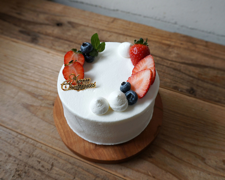 木製のテーブルの上に置かれた小さな円形の白いクリスマスケーキ。生クリームで覆われ、上部にはスライスされたイチゴ、ブルーベリー、ミントの葉、そして「Merry Christmas」と書かれた金色のピックで飾られている。