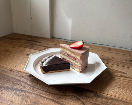 木のテーブルに置かれた白い八角形のプレート。瑞々しいいちごが乗った層の美しいショートケーキと、滑らかな質感のチョコレートタルトが並び、柔らかな日差しが差し込んでいる。