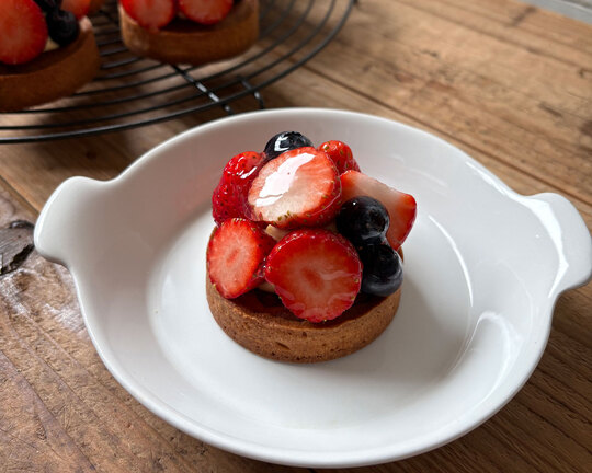 瑞々しいイチゴとブルーベリーがたっぷりと盛り付けられたイチゴタルト。白い持ち手付きの小皿に乗り、木のテーブルの上で自然光に照らされている。