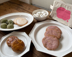 木の卓上に並ぶホワイトデーセット。数種類の焼き菓子、メレンゲ、ピンクのロゴ入りポーチが柔らかな光に包まれている。