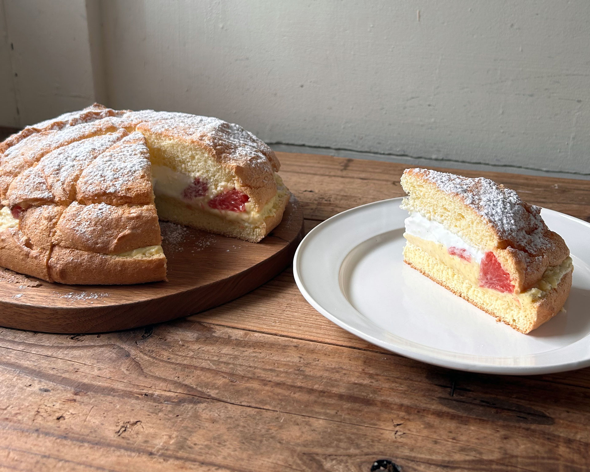 粉砂糖をかけたシュー生地のカスタードケーキ。中にカスタードクリームとピンクグレープフルーツが入っており、木製のまな板と白い皿に盛り付けられている。