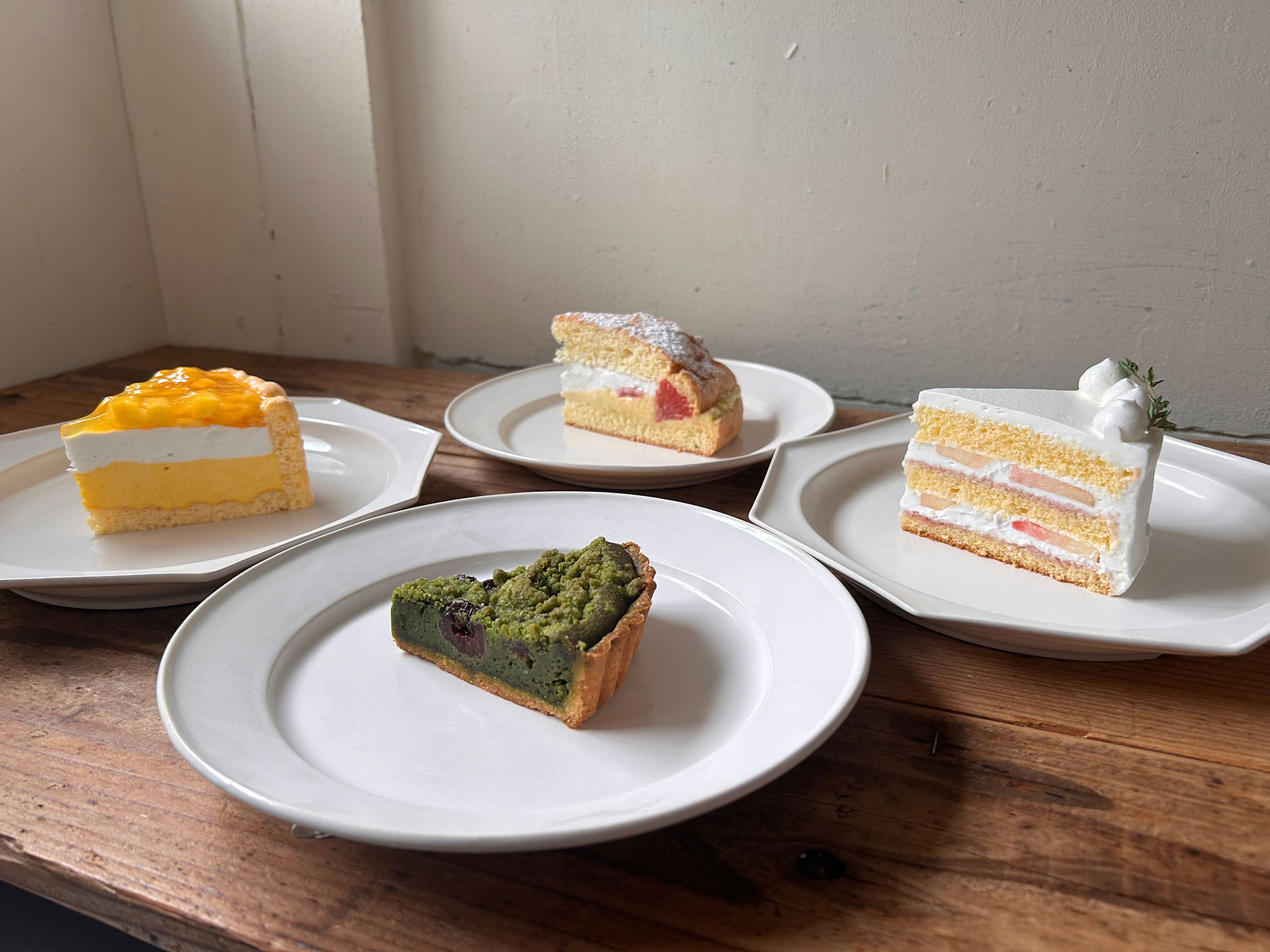 木製テーブルに並ぶマンゴーシャルロット、桃ショートケーキ、抹茶チェリークランブルタルト、カスタードケーキ